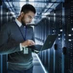 IT technician works on a laptop in big data centre full of rack servers.