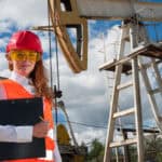 Engineer in the oilfield wearing red helmet and work clothes, with pumpjack and wellhead in the background.