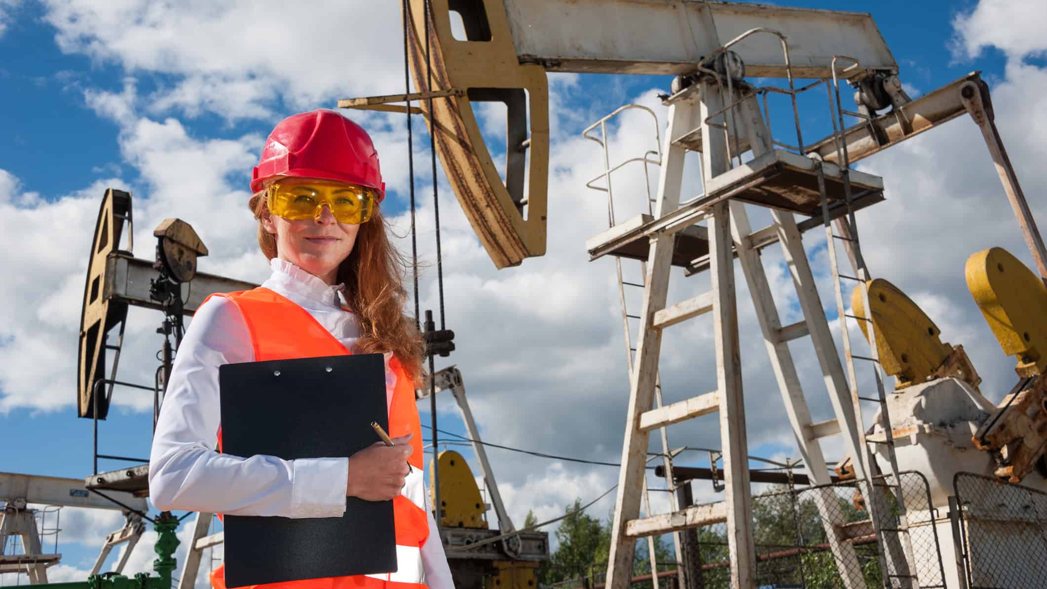 Engineer in the oilfield wearing red helmet and work clothes, with pumpjack and wellhead in the background.