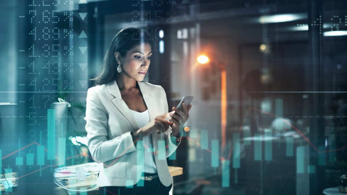 Shot of a young businesswoman using her phone at work, with stock market related images in the background.
