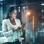 Shot of a young businesswoman using her phone at work, with stock market related images in the background.