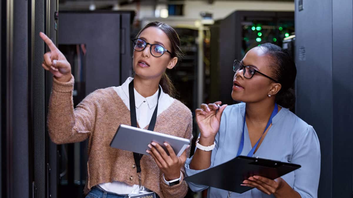 Two woman holding a tablet and clipboard examine a server rack in a data centre.