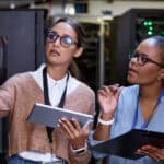 Two woman holding a tablet and clipboard examine a server rack in a data centre.