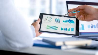 Two business men looking at financial data, charts, and graphs on a digital tablet and a laptop computer.