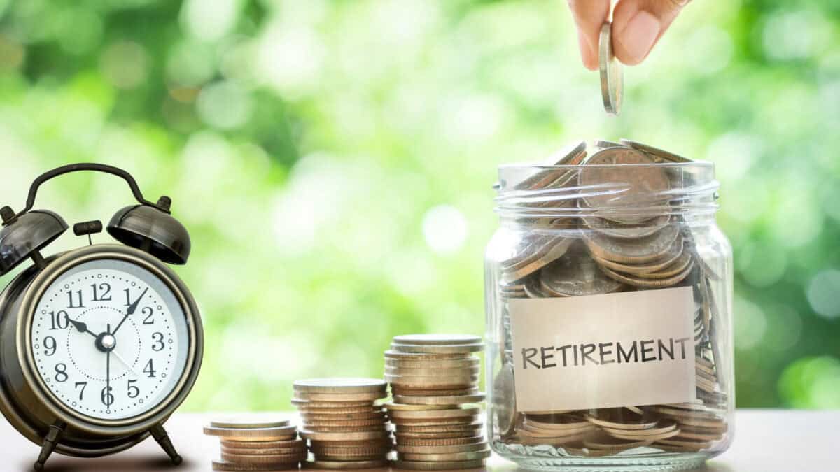 Hand putting coins in a glass jar that says retirement, with a retro alarm clock on the other side, and piles of increasing coins in the middle.