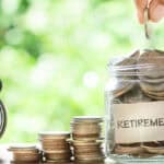 Hand putting coins in a glass jar that says retirement, with a retro alarm clock on the other side, and piles of increasing coins in the middle.