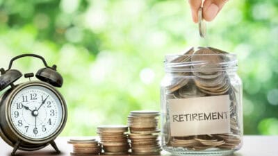 Hand putting coins in a glass jar that says retirement, with a retro alarm clock on the other side, and piles of increasing coins in the middle.