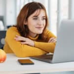 Businesswoman with a pleased smile reading on her laptop at a desk in the office with a look of satisfaction.