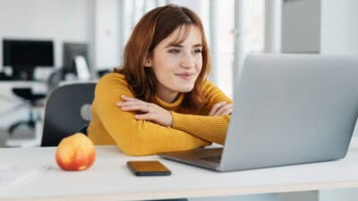 Businesswoman with a pleased smile reading on her laptop at a desk in the office with a look of satisfaction.