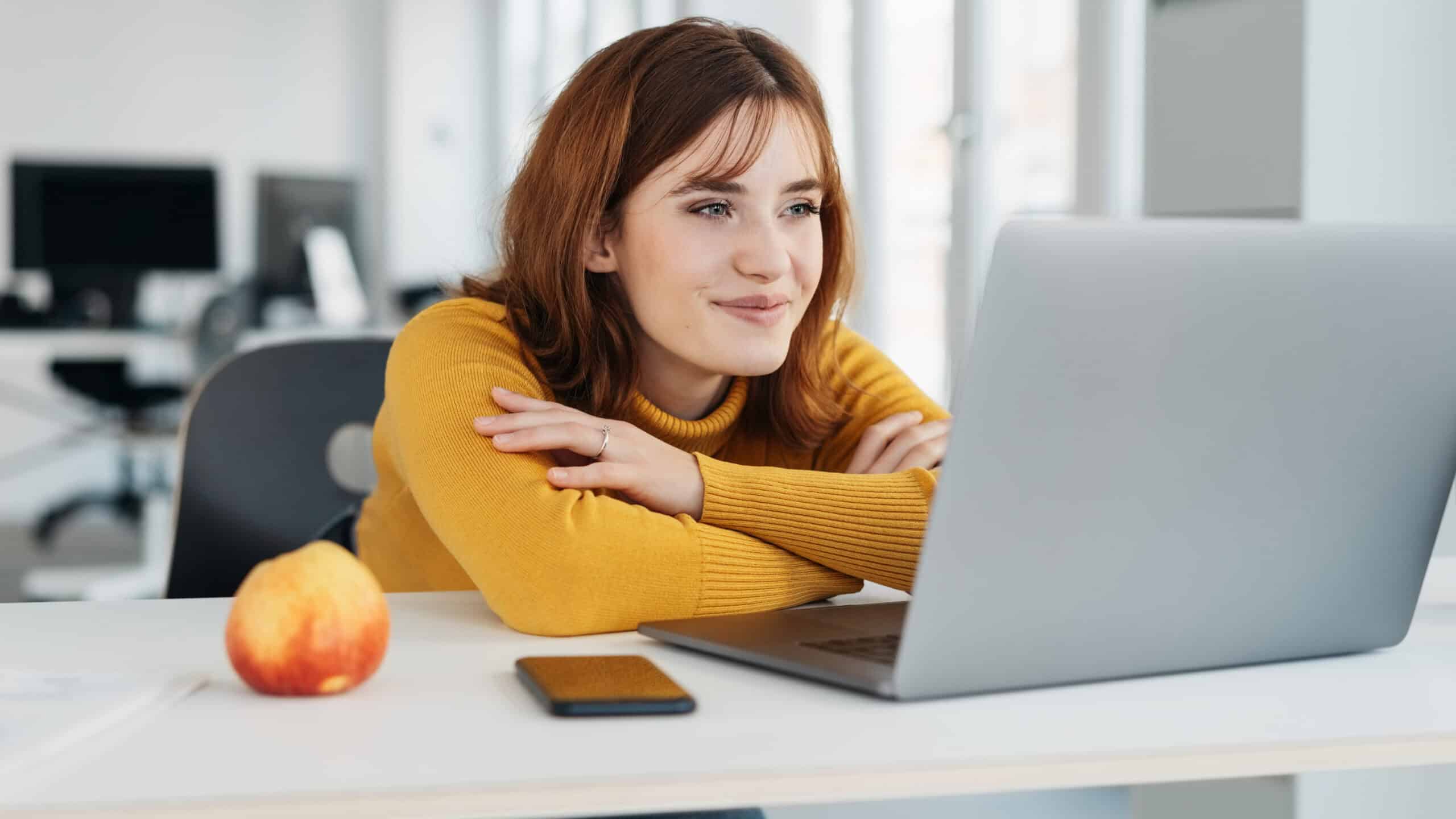Businesswoman with a pleased smile reading on her laptop at a desk in the office with a look of satisfaction.