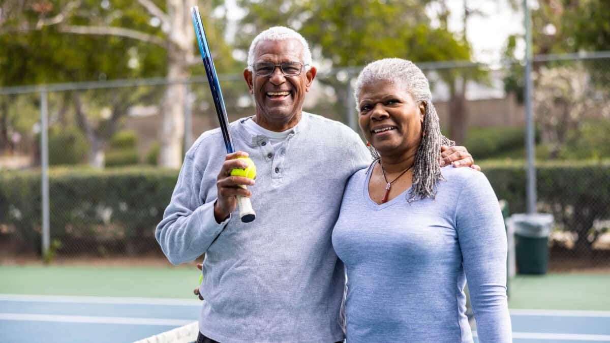 Couple posing for photo at a tennis court, with man holding a racquet and ball.