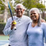 Couple posing for photo at a tennis court, with man holding a racquet and ball.