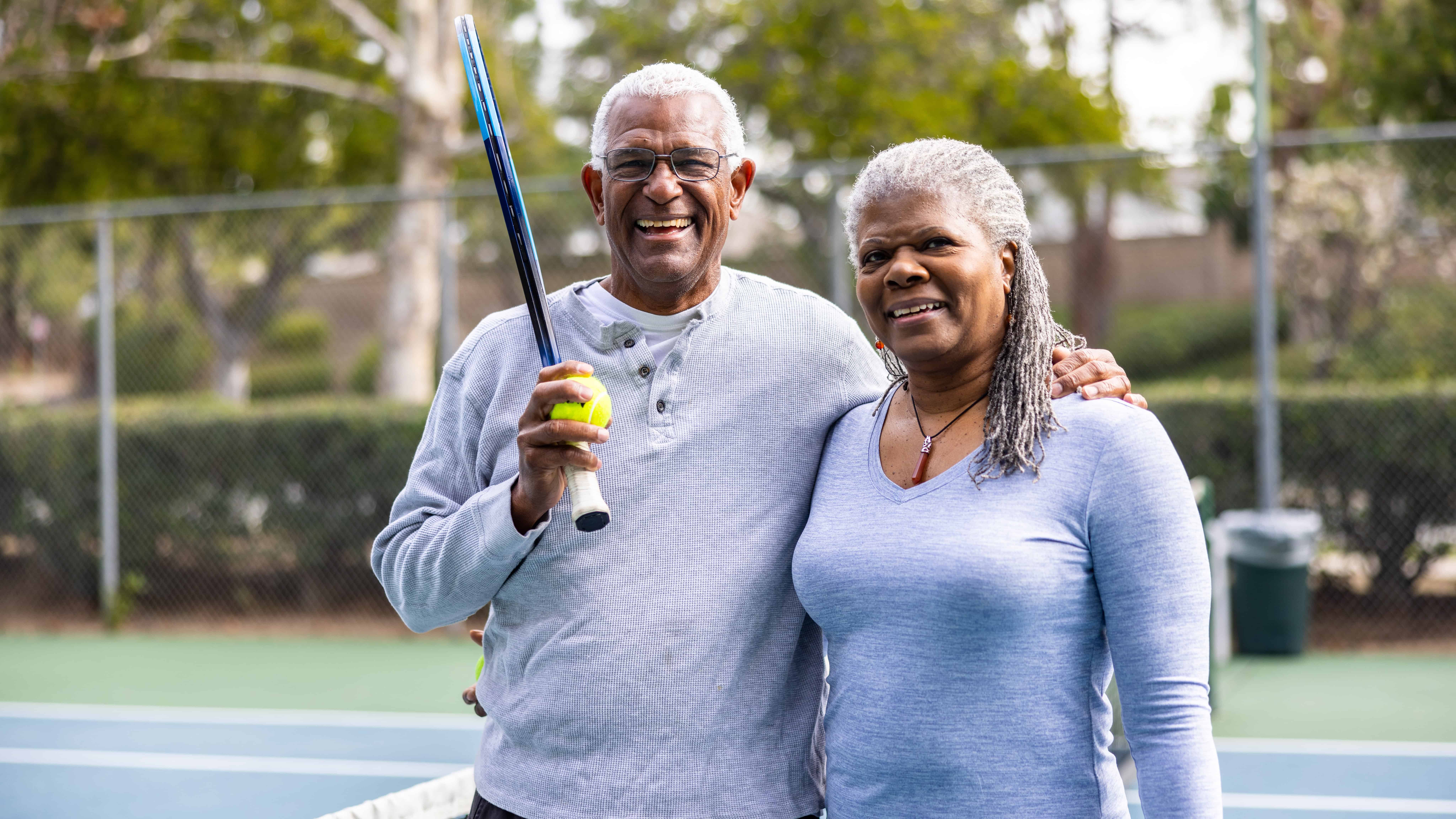 Couple posing for photo at a tennis court, with man holding a racquet and ball.