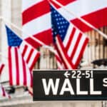 Wall Street sign with New York Stock Exchange building out of focus in the background with American flags.