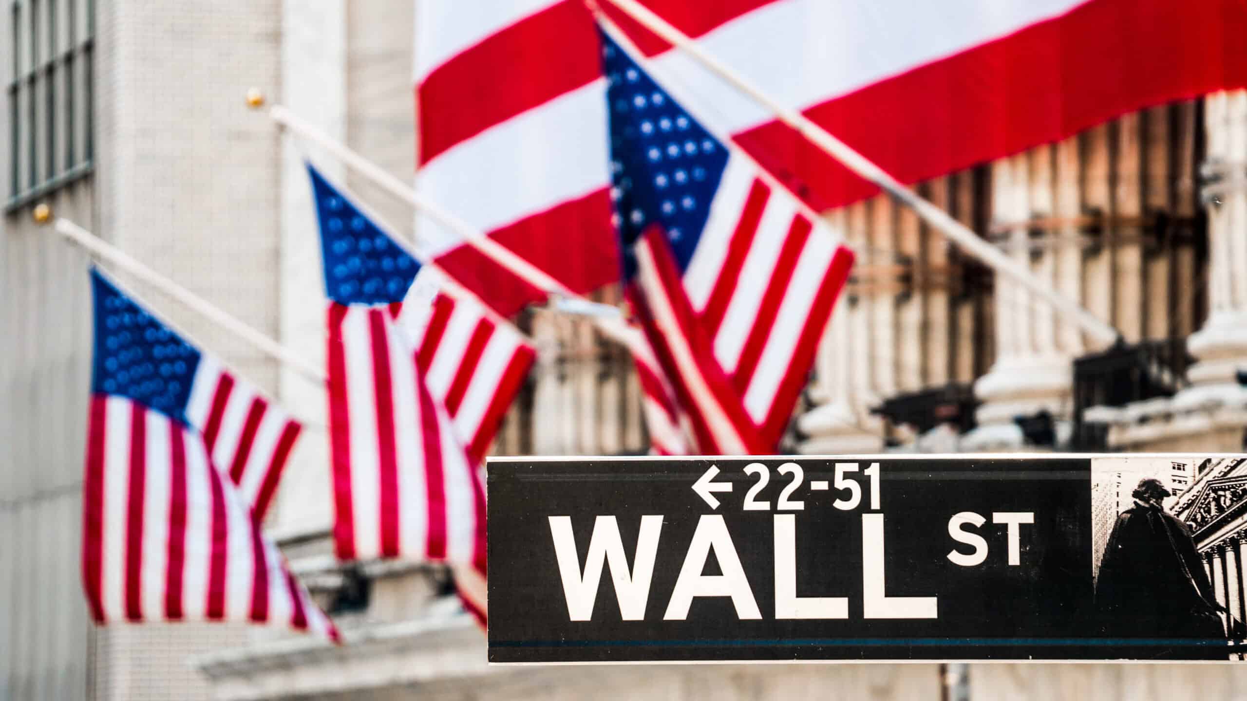 Wall Street sign with New York Stock Exchange building out of focus in the background with American flags.
