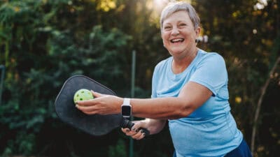 Happy and smiling retiree posing with pickleball and paddle.