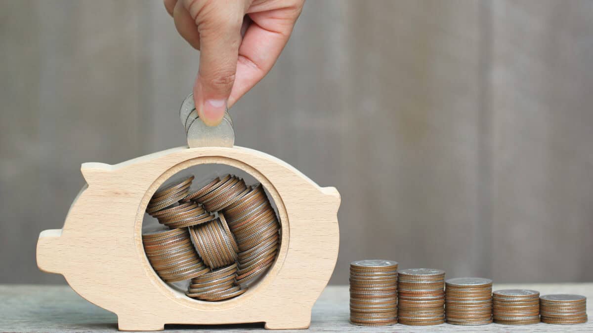 Man putting coins in a wooden piggy bank next to piles of coins.
