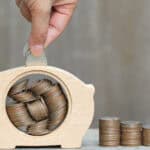 Man putting coins in a wooden piggy bank next to piles of coins.
