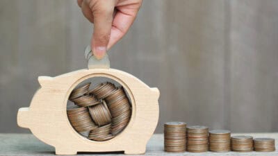 Man putting coins in a wooden piggy bank next to piles of coins.