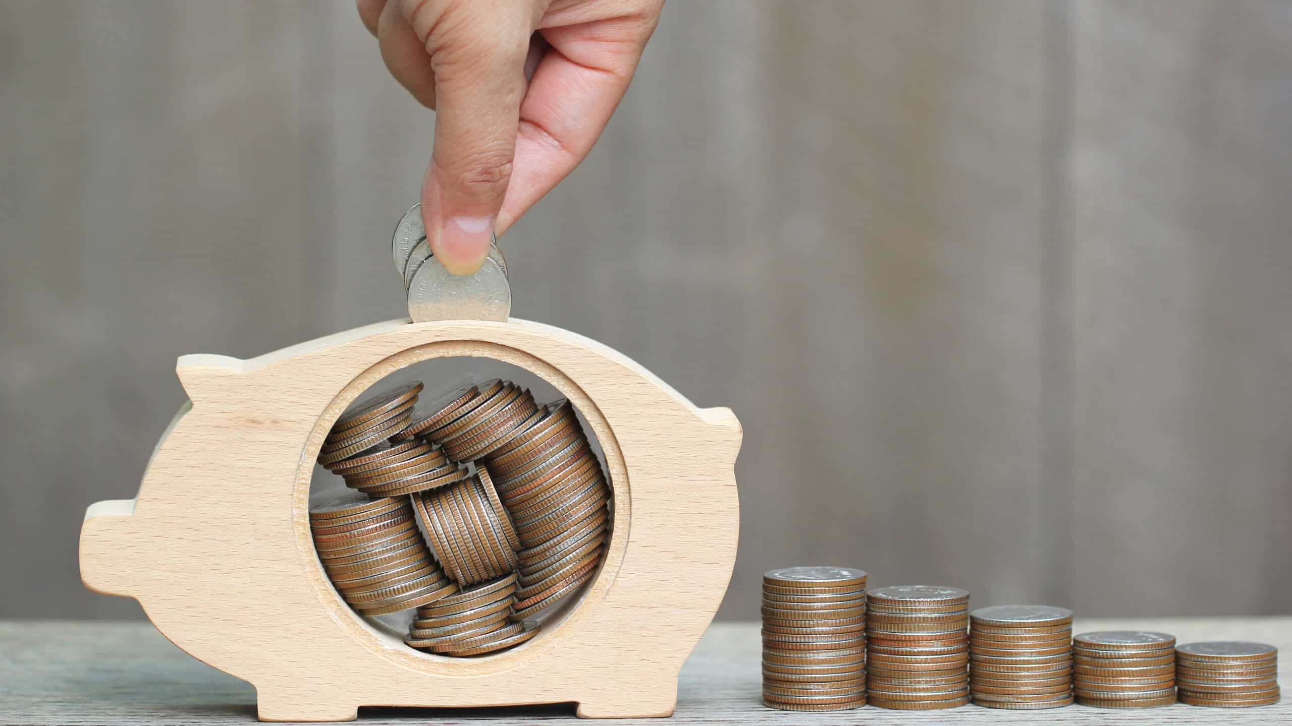 Man putting coins in a wooden piggy bank next to piles of coins.