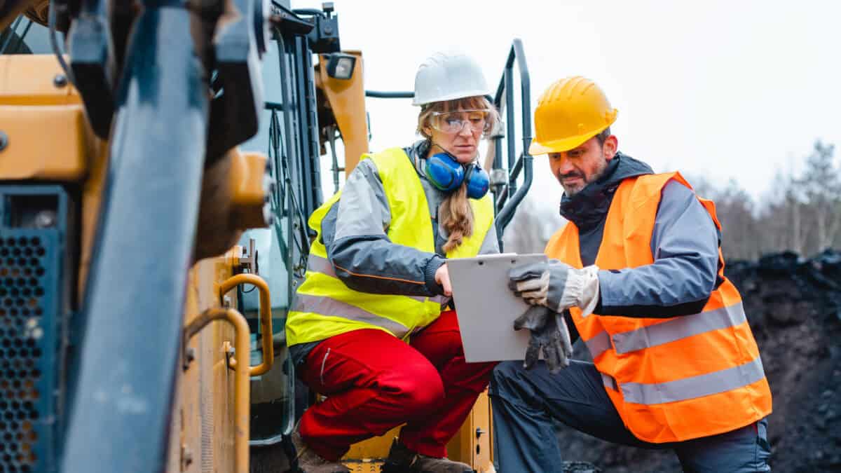 Woman and man worker in quarry on excavation machine looking at a clipboard.