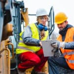 Woman and man worker in quarry on excavation machine looking at a clipboard.