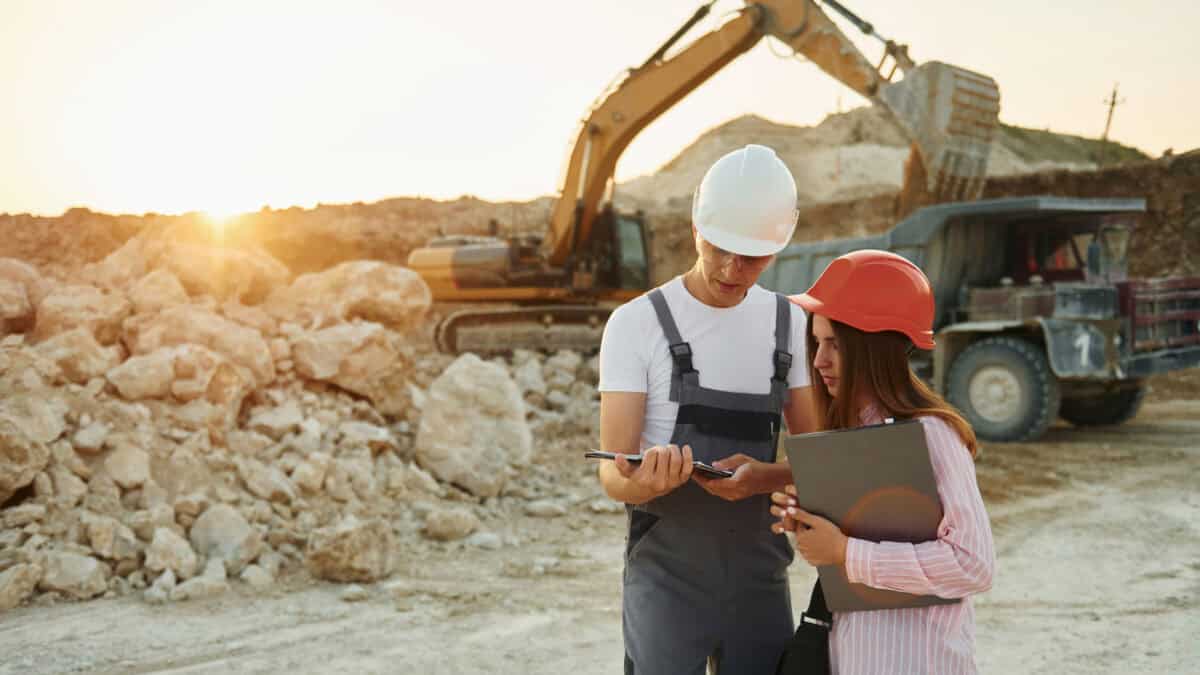 Two workers on a tablet at a mine site, with mining machinery behind them.