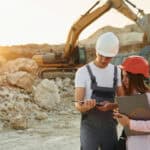 Two workers on a tablet at a mine site, with mining machinery behind them.
