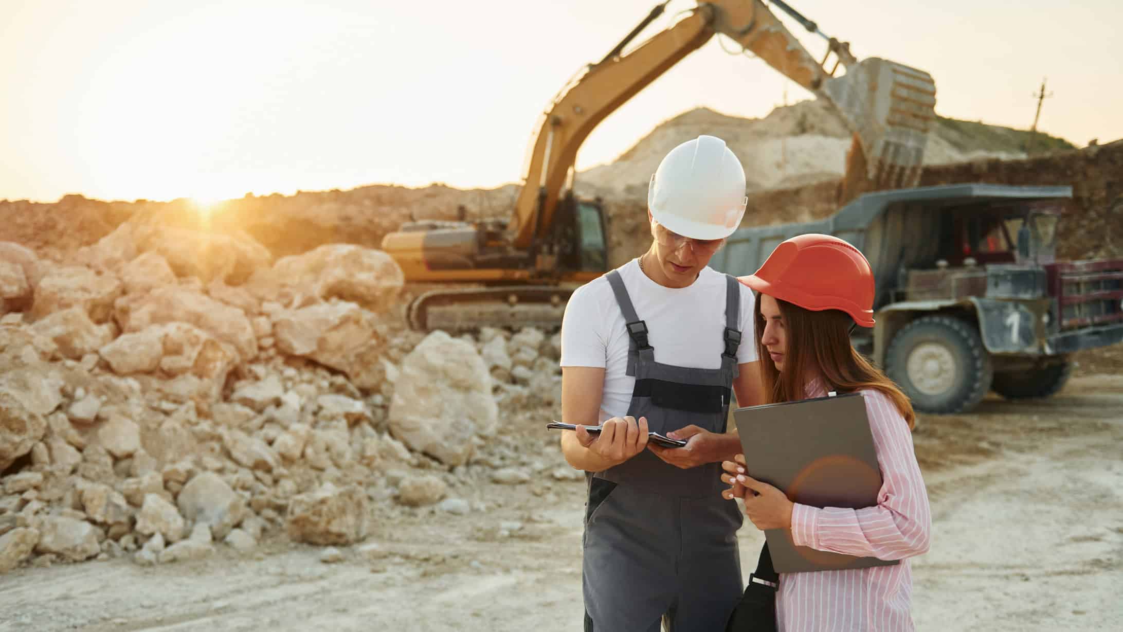 Two workers on a tablet at a mine site, with mining machinery behind them.