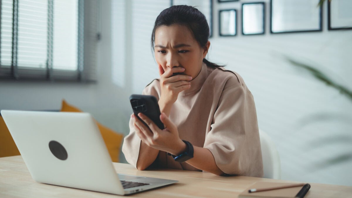 Worried woman with hand on her mouth looks at her phone with a laptop open in front of her.