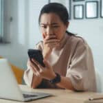 Worried woman with hand on her mouth looks at her phone with a laptop open in front of her.