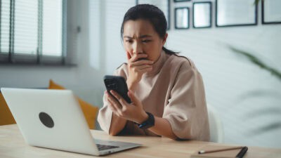 Worried woman with hand on her mouth looks at her phone with a laptop open in front of her.