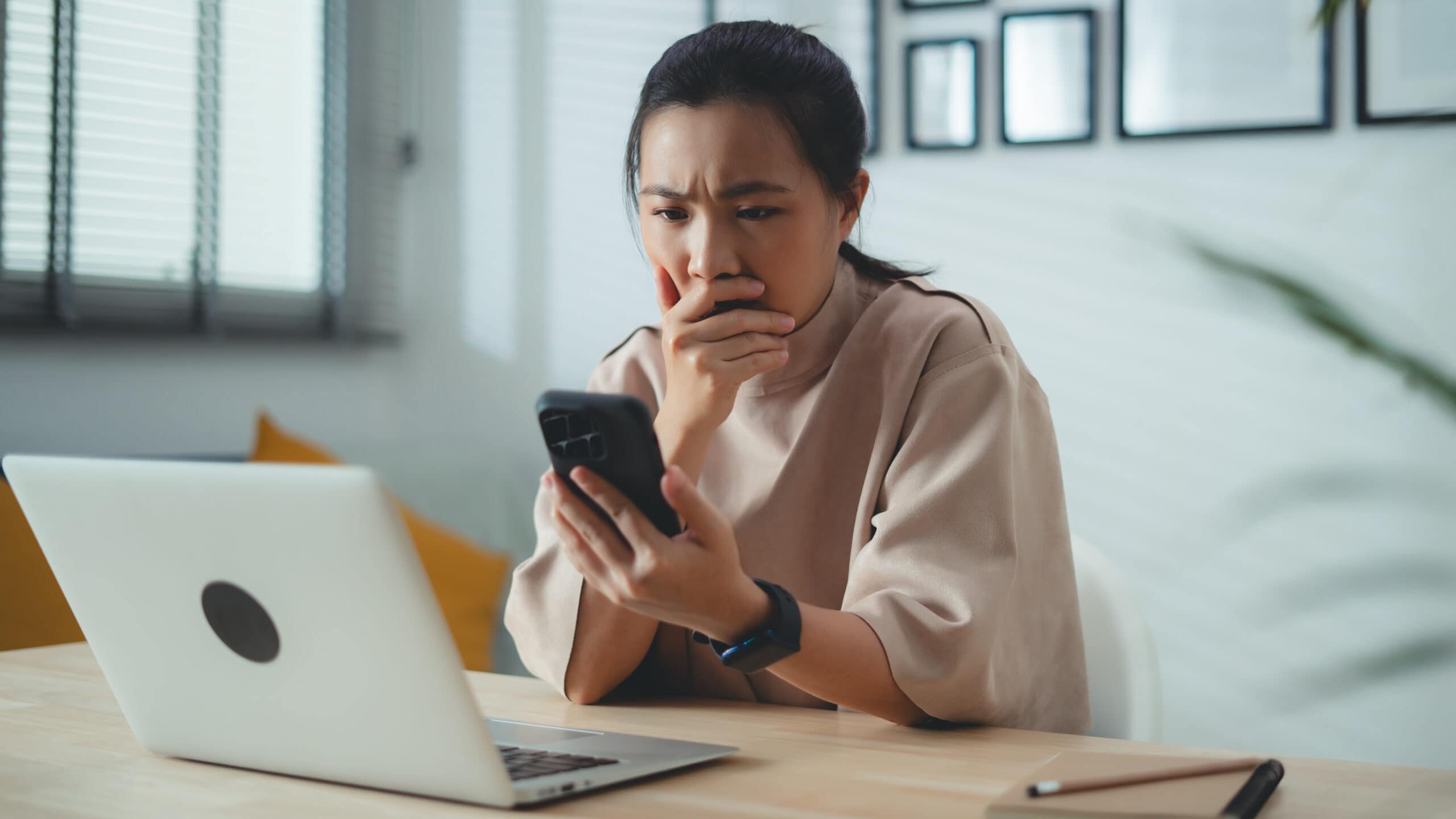 Worried woman with hand on her mouth looks at her phone with a laptop open in front of her.