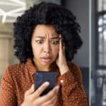 A distressed young woman reads bad news on her smartphone while standing in a modern indoor setting.