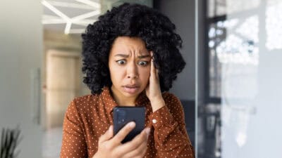 A distressed young woman reads bad news on her smartphone while standing in a modern indoor setting.