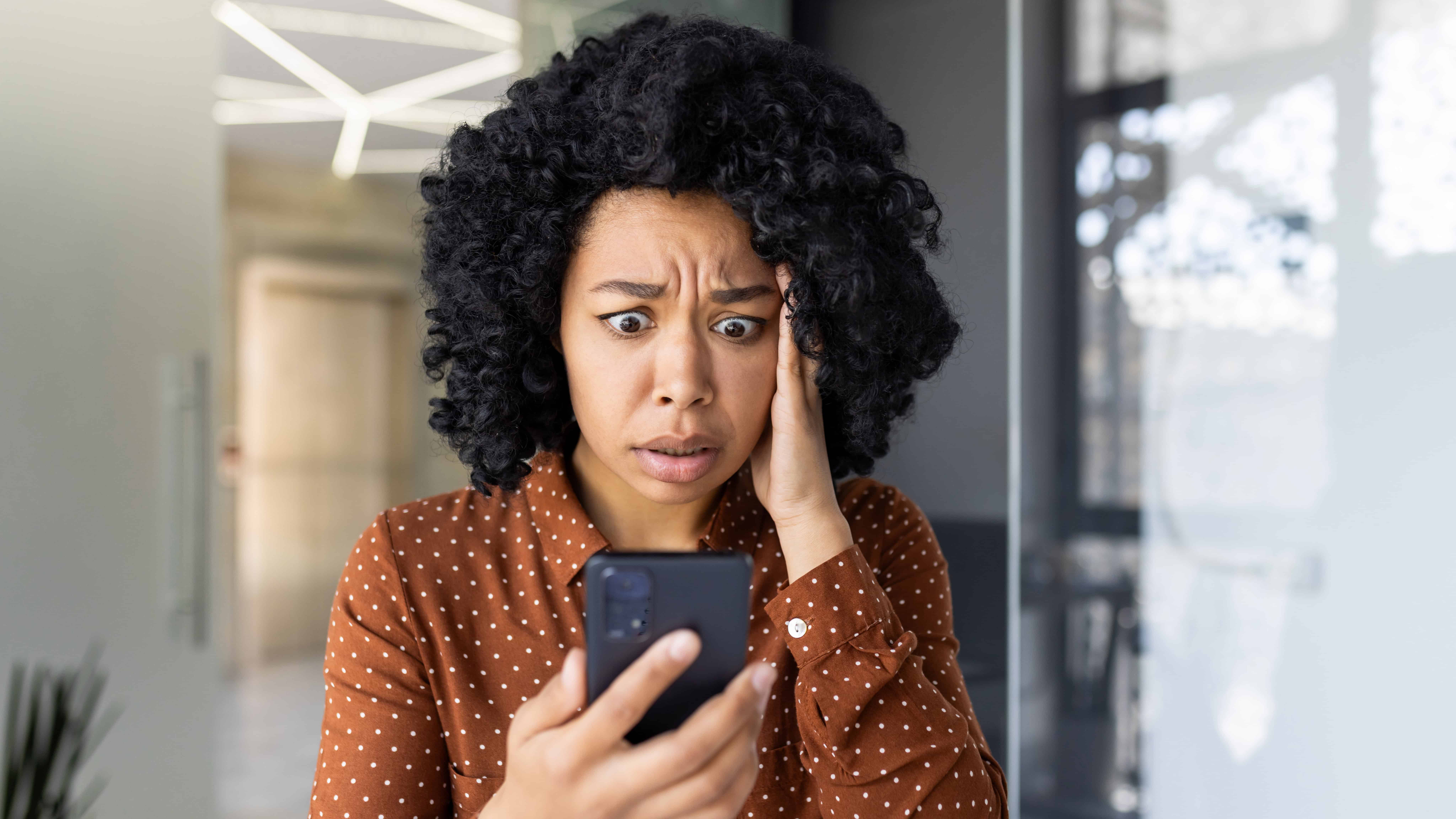 A distressed young woman reads bad news on her smartphone while standing in a modern indoor setting.