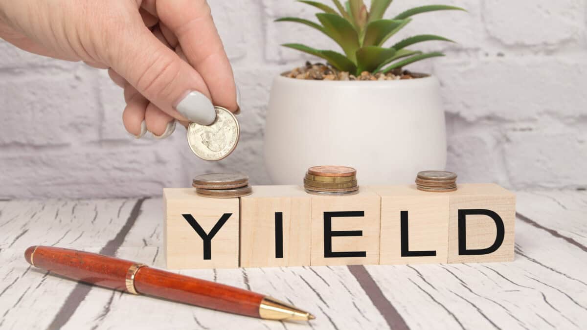Yield written on wooden blocks with a hand putting coins on top, with a plant and pen on the table.