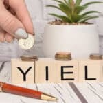 Yield written on wooden blocks with a hand putting coins on top, with a plant and pen on the table.