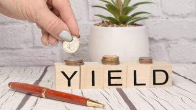 Yield written on wooden blocks with a hand putting coins on top, with a plant and pen on the table.