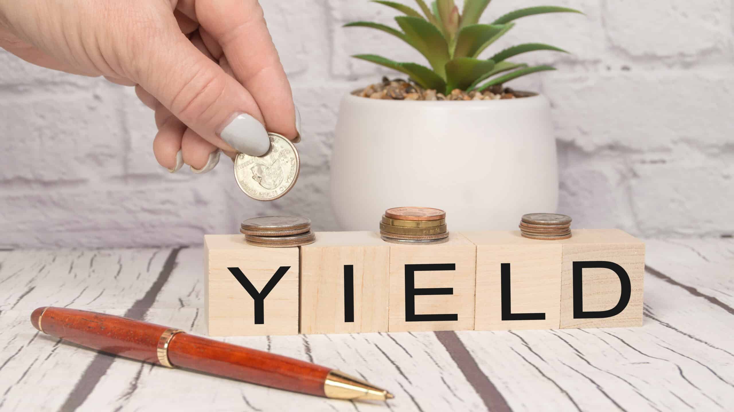 Yield written on wooden blocks with a hand putting coins on top, with a plant and pen on the table.