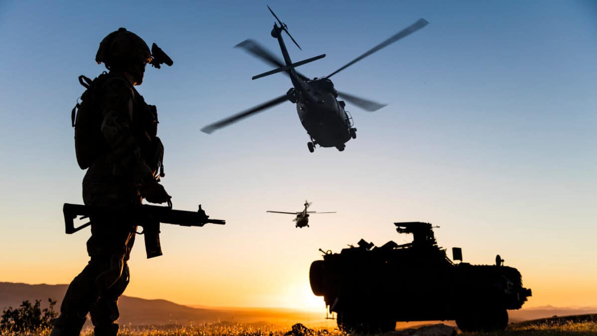Man in army uniform holding a gun with two helicopters in the sky and a defence vehicle on the ground.
