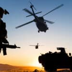Man in army uniform holding a gun with two helicopters in the sky and a defence vehicle on the ground.
