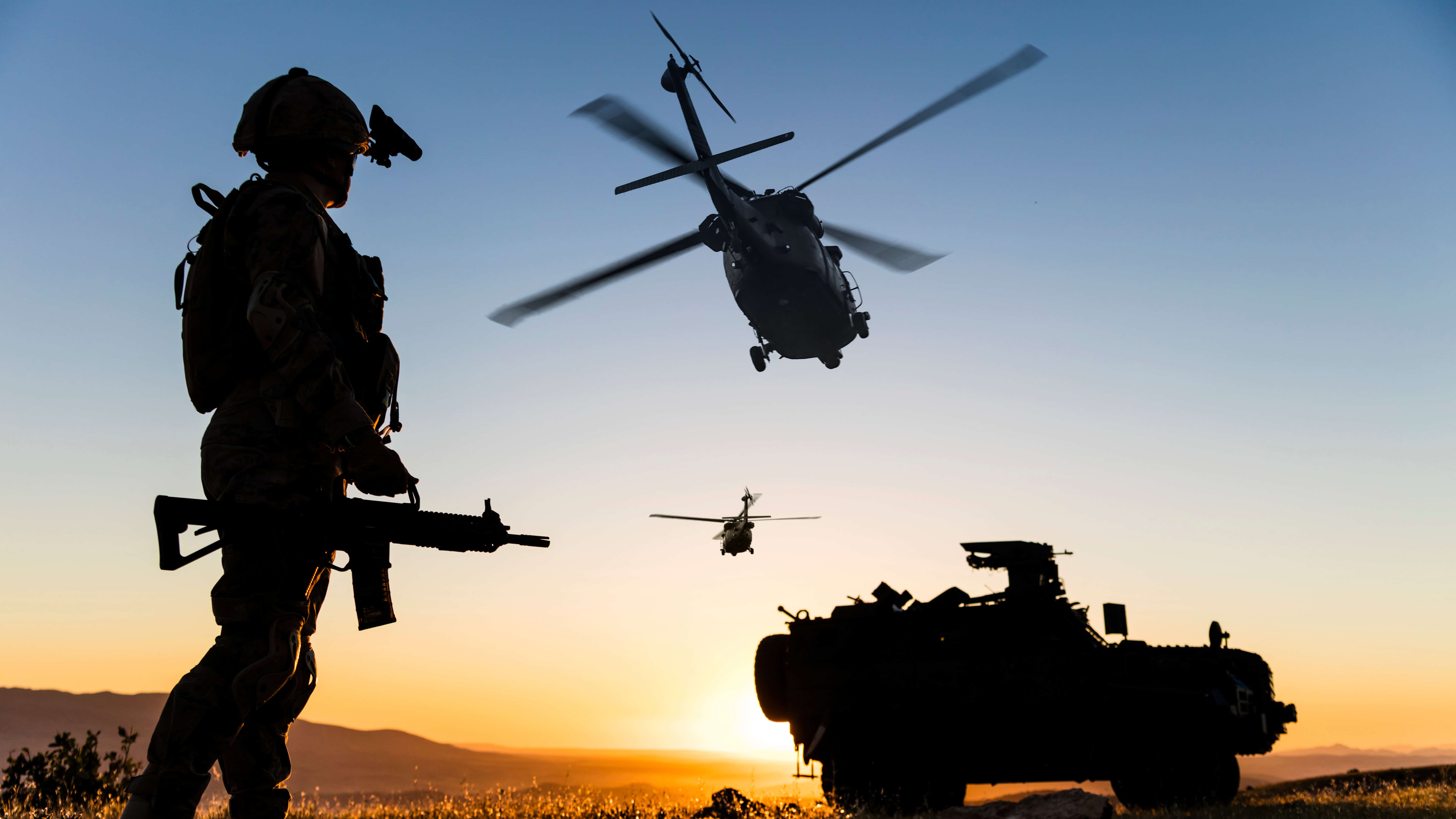 Man in army uniform holding a gun with two helicopters in the sky and a defence vehicle on the ground.