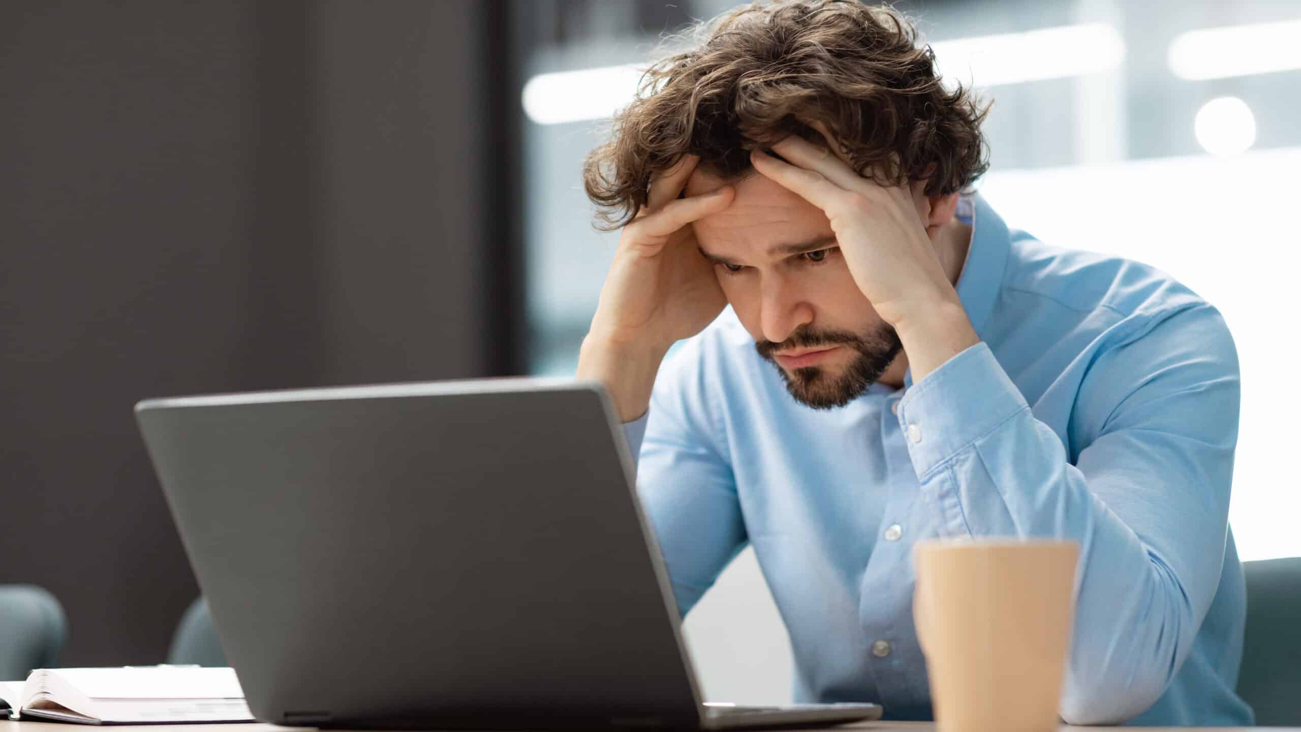 Sad man sitting at desk and grabbing his head as he looks at a laptop.