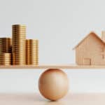 Wooden house and golden coins on balancing scale.