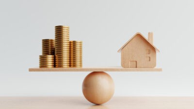 Wooden house and golden coins on balancing scale.