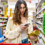 Woman using smartphone to check product details while shopping in a grocery store aisle.