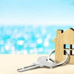Model of house and key on sandy beach with sea and sky in the background.