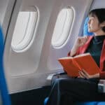 Woman looking through an airplane window while holding a book.