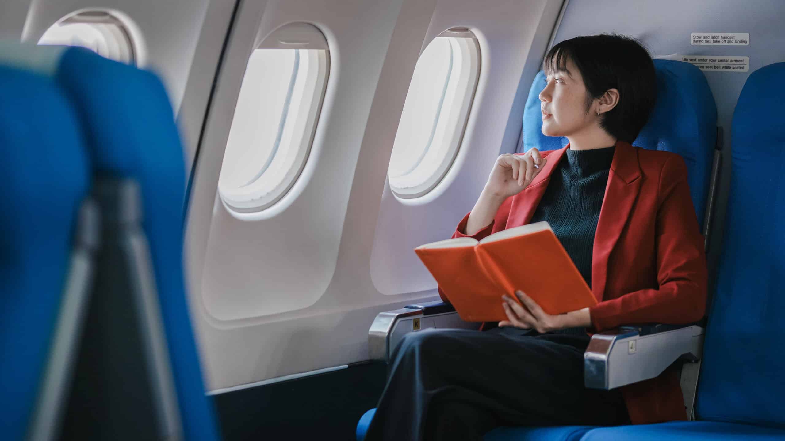 Woman looking through an airplane window while holding a book.
